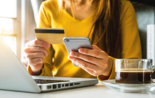 A woman sits at her computer with her phone and credit card in hand, ready to make a purchase online.