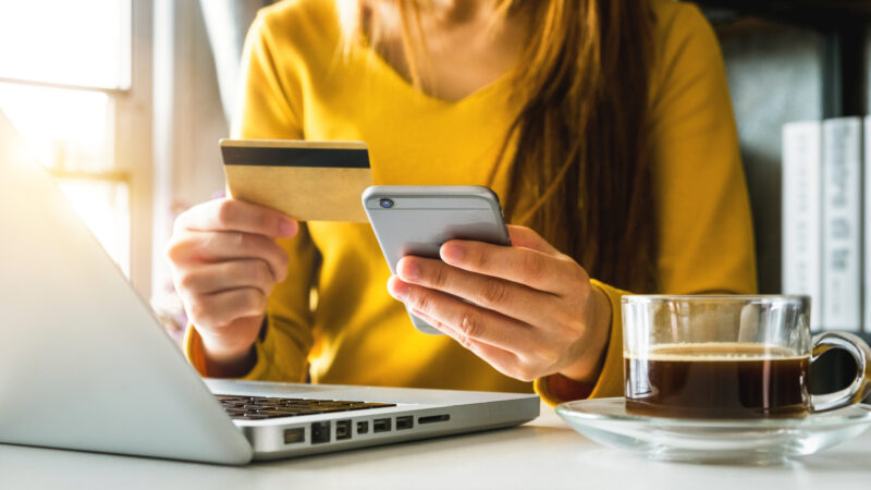 A woman sits at her computer with her phone and credit card in hand, ready to make a purchase online.